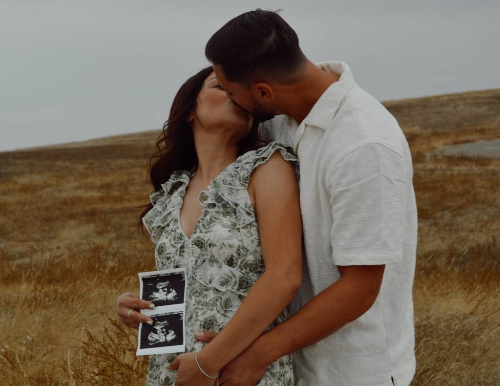 Expecting couple kissing in a golden field while holding ultrasound photos
